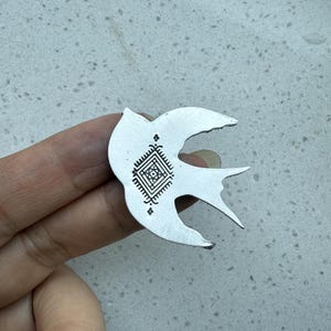 May include: A silver-colored bird-shaped pendant with a geometric design on its body. The pendant has a detailed, etched pattern within a square shape, and is held against a light-colored surface.