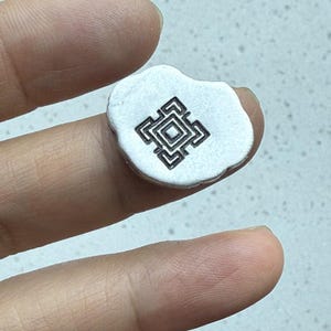 May include: A small, irregularly shaped, silver-colored charm with a stamped geometric design in black. The charm is held between fingers, showcasing its size and detail. The design is a square within a square, with additional geometric shapes.