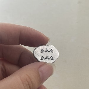 May include: A silver metal charm with a design of three rows of small black dots arranged in a triangular pattern.