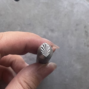May include: A close-up of a metal stamp with a geometric leaf design. The stamp is held in a hand, and the design is visible on the top of the stamp.