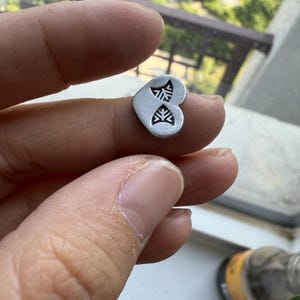 May include: A small, silver-colored heart-shaped charm with two stylized leaf designs. The charm is held between fingers, showcasing its size and detail. The background is blurred, focusing attention on the jewelry.