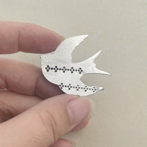 May include: A silver metal bird pendant with a dotted design. The bird is in flight with its wings spread out.