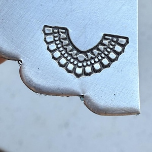 May include: A close-up of a metal stamp with a design of a decorative collar or necklace. The stamp is silver in color and has a detailed design.
