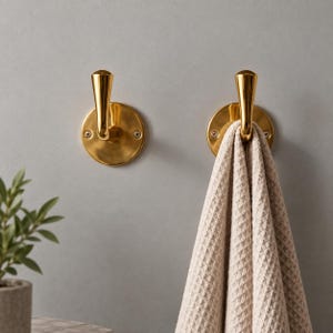 May include: Two polished brass wall hooks with circular mounting plates and vertical stems. One hook is empty, while the other holds a beige waffle-textured towel draped over its curved top. The hardware is secured with visible screws.