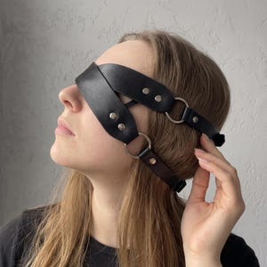 Può includere: Una benda nera in pelle con due cinturini sovrapposti che coprono gli occhi. Il design include anelli in metallo argentato sui lati e rivetti argentati che fissano i cinturini, creando un accessorio per la testa strutturato e regolabile.