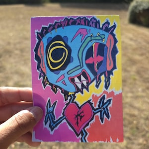 May include: A rectangular sticker featuring a vibrant, multi-colored illustration of a stylized creature with a blue face, a large yellow spiral eye, and sharp teeth. A red heart with a crack in the center sits below the head, surrounded by pink, yellow, and orange background sections with bold black outlines.