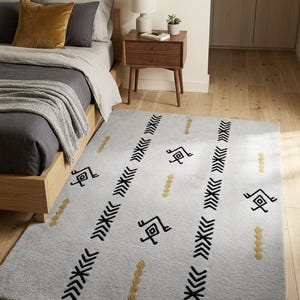Könnte beinhalten: Ein rechteckiger Teppich mit hellgrauer Grundfläche und zwei markanten vertikalen Streifen aus schwarzen Chevron-Mustern. Das Design enthält verstreute goldfarbene kreisförmige Akzente und kleine schwarze geometrische Symbole zwischen den Streifen.