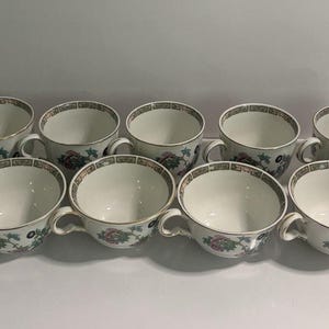 May include: A set of eleven white ceramic teacups arranged in two rows. Each cup features a delicate floral pattern in shades of pink, green, and blue, with a decorative geometric border along the rim and a gold-toned handle and trim.