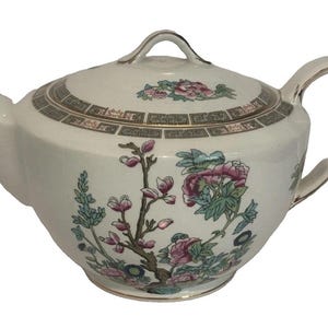 May include: A white ceramic teapot featuring a floral pattern with pink and green blossoms on the body. The lid and rim are decorated with a geometric border in brown and gold. The handle, spout, and lid knob are accented with a metallic gold trim.