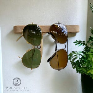 Può includere: Un supporto da parete in legno chiaro che sostiene due paia di occhiali da sole in stile aviatore. La coppia a sinistra presenta montature dorate con lenti color verde, mentre la coppia a destra ha montature scure con lenti color marrone.