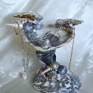 May include: A decorative jewellery holder crafted from natural seashells, featuring a tiered design with a wide base and a bowl-like top. The structure is adorned with various small spiral shells and white pearls, holding silver and gold-toned chains.