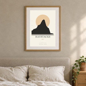 Peut inclure: Une affiche encadrée représentant la silhouette noire minimaliste de la montagne Haystacks devant un grand soleil beige. Le texte indique HAYSTACKS, 597 m, ainsi que les coordonnées du Lake District en Angleterre sur un fond crème avec un cadre en bois.