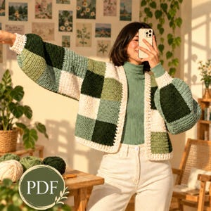 May include: A crochet cardigan featuring a patchwork design of square blocks in shades of dark green, sage green, and cream. The garment has long, voluminous sleeves with ribbed cuffs and an open front, worn over a sage green turtleneck shirt.
