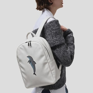 Puede incluir: Una mochila blanca con una ilustración gráfica de un delfín gris y azul en el panel frontal. La mochila cuenta con un asa superior, correas ajustables para los hombros y un compartimento principal con cremallera y tiradores negros.