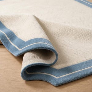May include: A rectangular area rug featuring a cream-coloured centre with a subtle, raised linear texture. The rug is bordered by a wide, solid blue band and a thin, cream-coloured inner stripe. The corner of the rug is folded over to reveal the backing.