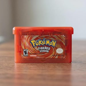 Puede incluir: Un cartucho de videojuego Game Boy Advance de color naranja translúcido para Pokémon FireRed Version. La etiqueta presenta un fondo rojo y amarillo con el logotipo de Pokémon, el texto FireRed Version, una clasificación ESRB E y la marca Nintendo.