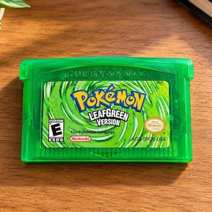 May include: A translucent green Game Boy Advance video game cartridge for Pokémon LeafGreen Version. The label features a swirling green background, the Pokémon logo, the ESRB E rating, and the Nintendo Official Seal of Quality text.
