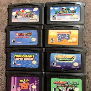 Può includere: Una collezione di otto cartucce per videogiochi Game Boy Advance nere disposte in due colonne. Le etichette presentano titoli tra cui Super Mario Advance, Super Mario World, Yoshi's Island, Super Mario Bros 3, Mario Kart, Mario Party, Mario vs Donkey Kong e Mario and Luigi Superstar Saga.