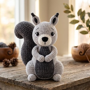 Puede incluir: Un peluche de ardilla tejido a ganchillo con cabeza y cuerpo de color gris claro, pecho, orejas y una gran cola tupida de color gris oscuro. La ardilla tiene ojos de seguridad negros, nariz negra bordada y una boca pequeña de hilo.