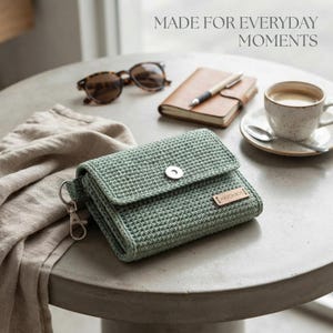 May include: A sage green crocheted wallet featuring a silver-tone snap closure, a metal lobster clasp on the side, and a small rectangular tag labeled "HANDMADE". The text "MADE FOR EVERYDAY MOMENTS" is visible in the upper right corner.