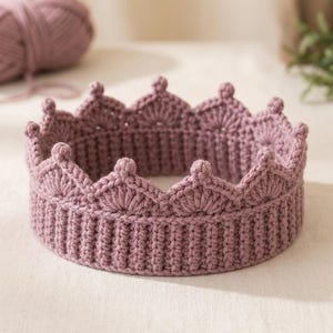 May include: A dusty rose crochet crown featuring a ribbed base and a scalloped top edge with small, rounded points. The crown is constructed from yarn and displays a consistent, textured stitch pattern throughout its circular design.