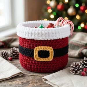 May include: A cylindrical crochet basket designed to resemble a Santa Claus suit, featuring a deep red body, a thick white top rim, a black belt band, and a yellow rectangular buckle. The basket is filled with candy canes and small ornaments.