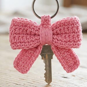May include: A pink crocheted bow keyring featuring a textured yarn construction with a central wrapped knot detail. A silver metal split ring is attached to the top of the bow, and a standard silver metal house key is inserted into the centre.