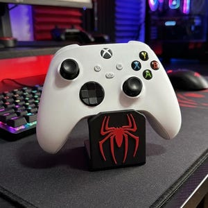 Peut inclure: Une manette de jeu vidéo sans fil blanche avec des joysticks noirs et une croix directionnelle, posée sur un support rectangulaire noir arborant un emblème d'araignée rouge bien visible sur la face avant, pour les consoles de jeu.