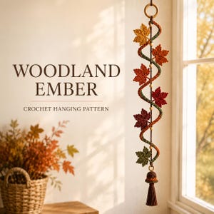 Op de afbeelding: Een gehaakte hangdecoratie met een spiraalvormig koord versierd met herfstkleurige esdoornbladeren in geel, oranje, rood en groen, geaccentueerd met houten kralen, een bovenring en een onderste kwastje. Tekst: WOODLAND EMBER.