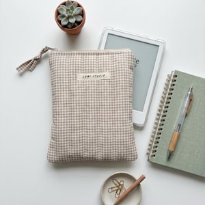 May include: A rectangular, quilted zipper pouch featuring a beige and white gingham pattern. A small fabric label on the front displays the text "LUMI STUDIO". The pouch includes a matching gingham fabric zipper pull attached to a silver metal zipper.