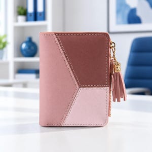 May include: A bi-fold wallet featuring a geometric color-blocked design in shades of dusty rose, mauve, and light pink. The wallet includes a gold-toned metal zipper closure on the side, accented with a matching pink leather tassel charm.