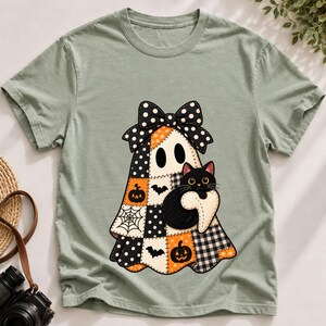 Può includere: Una t-shirt verde salvia con una grafica centrale raffigurante un fantasma che indossa un grande fiocco a pois bianchi e neri. Il fantasma presenta un motivo patchwork di zucche arancioni, pipistrelli neri, ragnatele e quadretti.