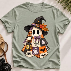 May include: A sage green t-shirt featuring a central graphic of a ghost wearing a black witch hat adorned with orange flowers and leaves. The ghost has a patchwork body pattern with small ghosts, bats, spiders, and a lantern illustration.