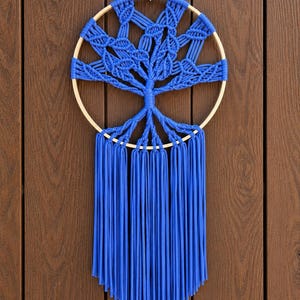 Op de afbeelding: Een wandhanger bestaande uit een ronde houten ring met een blauw macraméontwerp in de vorm van een boom. Het bovenste gedeelte toont ingewikkelde geknoopte takken, terwijl het onderste gedeelte bestaat uit lange, rechte, blauwe franjes.