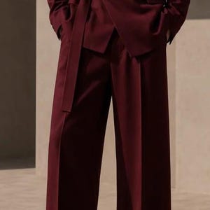 Puede incluir: Un traje de color burdeos profundo que consta de una americana y pantalones anchos. La americana presenta un estilo cruzado con un cinturón lateral y una hebilla metálica rectangular. Los pantalones tienen un corte recto con una abertura vertical.