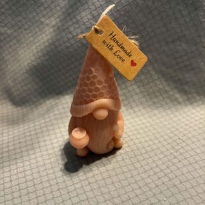 Peut inclure: Une bougie rose clair en forme de gnome avec un motif alvéolé sur le chapeau. Le gnome tient un petit objet rond dans une main et possède une longue barbe. Une étiquette en papier brun est attachée, portant l'inscription Handmade with Love.