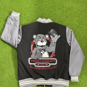 May include: A black and grey varsity jacket featuring a graphic of a bear character holding a microphone and wearing a top hat. The text reads "FREDDY FAZBEAR'S PIZZA" and "Where fantasy and fun come to life!" with red stars on a black background.