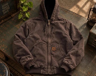 May include: A dark brown hooded jacket featuring a full-length front zip, a drawstring hood, ribbed cuffs and hem, and a front kangaroo pocket with a rectangular Carhartt brand patch stitched onto the lower left side of the pocket.