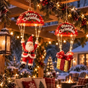 May include: Two hanging decorations featuring red umbrellas with the text Merry Christmas in white letters. One umbrella suspends a Santa Claus figure in a red suit, while the other suspends a gold gift box with a large red bow, both illuminated.