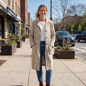 May include: A long, beige, button-down coat with a collar and two large front pockets, worn over a plain white t-shirt and blue denim jeans. The coat features a straight-cut silhouette, long sleeves, and a textured fabric finish, reaching mid-calf.