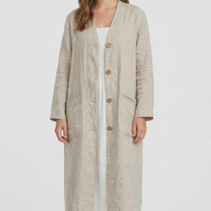 Puede incluir: Un abrigo tipo duster de lino beige de manga larga con escote en V, cuatro botones grandes de color marrón en la parte delantera y dos bolsillos de parche profundos en los laterales. El abrigo se lleva sobre un vestido blanco sencillo.