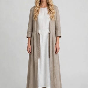Puede incluir: Un abrigo largo tipo duster de color beige, abierto por delante y con mangas tres cuartos, sobre un vestido blanco sencillo sin mangas hasta el suelo. El abrigo incluye un detalle de lazo en la cintura, bajo recto y pantalones anchos beige.