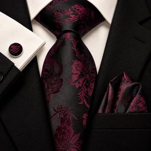 May include: A black necktie and matching pocket square featuring a dark burgundy floral pattern, worn with a white dress shirt, a black suit jacket, and a circular cufflink with a matching burgundy and black floral design on the shirt cuff.