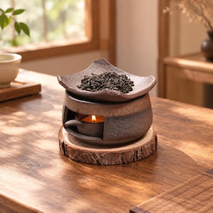 May include: A dark brown ceramic tea warmer with a textured surface and a square top tray holding loose tea leaves. A small lit candle sits inside the hollow base, which rests on a circular wooden slice with visible bark edges.