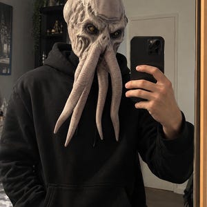 Op de afbeelding: Een masker voor het hele hoofd van een wezen met een bleke, getextureerde huid met diepe rimpels, opvallende tentakels die vanaf het gezicht naar beneden hangen en doordringende gele ogen. Het masker wordt gedragen met een zwarte hoodie.
