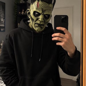 Puede incluir: Una máscara de monstruo de Frankenstein de cabeza completa con piel verde texturizada, parte superior negra plana, una cicatriz prominente en la frente y pernos plateados en el cuello, junto a una sudadera negra con capucha y bolsillo frontal.