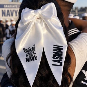 May include: A white satin hair bow featuring a black printed Navy eagle emblem on one tail and the name LASMAN in black block lettering on the other tail. The bow is secured in dark wavy hair, with long pointed tails hanging down the back.