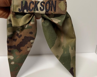 May include: A camouflage fabric hair bow featuring a central horizontal band embroidered with the name JACKSON in dark thread. The bow has two long, pointed tails and is constructed from a green, brown, and tan patterned textile material.
