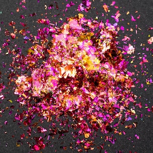 May include: A collection of small, irregular metallic flakes in vibrant shades of magenta, hot pink, and gold. These thin, reflective pieces are scattered in a dense cluster, creating a textured, shimmering effect with varied light reflections.
