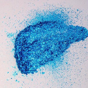 May include: A concentrated pile of vibrant blue metallic flakes with a reflective, crystalline texture. The particles vary in size, ranging from fine dust to larger, jagged shards, creating a dense, shimmering cluster of bright cyan and deep azure.